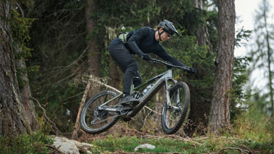 Je ziet een man op een grijze Cube Stereo Hybrid e-bike over een helling in het bos springen.