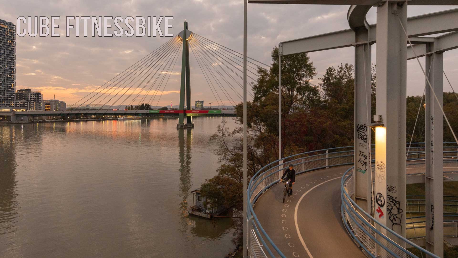 Je ziet een stad aan de rivier, op de voorgrond rijdt een fietser over het fietspad in de schemering. De foto is gelabeld met de titel Cube Fitnessbike.