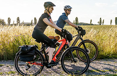 Je ziet een stel op e-bikes over een grindpad rijden. Er is hoog gras op de achtergrond.