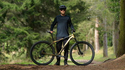 Je ziet een man achter zijn gouden Cube mountainbike op een bospad.