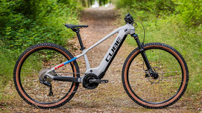 Je ziet een witte Cube e-bike voor kinderen met kleine details in blauw en rood. Hij staat op een overwoekerd bospad.