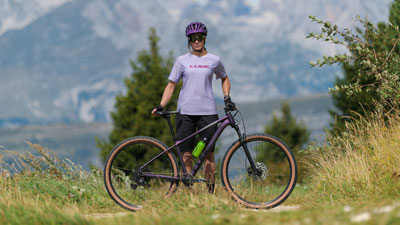 Je ziet een vrouw in paarse fietskleding, voor haar staat een paarse Cube mountainbike. Op de achtergrond is een bergachtig landschap te zien.