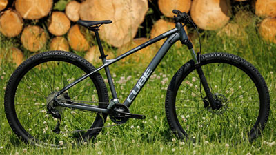 Je ziet een grijze Cube mountainbike in een weiland staan. Op de achtergrond ligt hout opgestapeld.