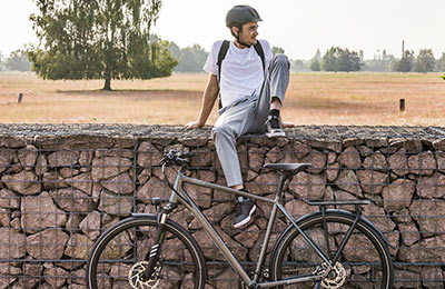 Een man met een helm zit op een muur. Op de voorgrond staat een grijze fiets en op de achtergrond een akkerlandschap.