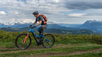 Je ziet een man op een blauwe Cube Stereo Hybrid e-bike met oranje details. Hij rijdt over een wandelpad en op de achtergrond zijn bergen te zien.
