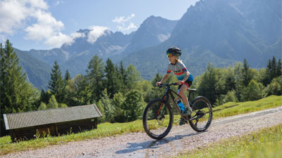 Een jongen rijdt op zijn mountainbike op een grindpad door een berglandschap. Op de achtergrond is een hut te zien.