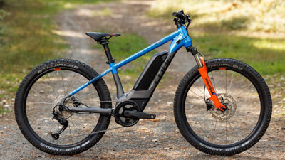 Je ziet een grijze Cube e-bike voor kinderen met blauwe en oranje details. Hij staat op een bospad.