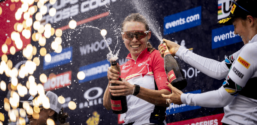 Kira Böhm viert haar overwinning in Canada op het podium door een geopende fles champagne te laten spuiten, naast een andere vrouwelijke deelnemer.